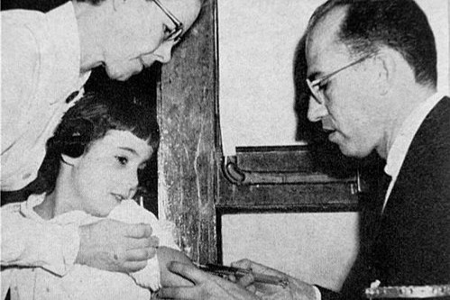 Dr. Jonas E. Salk administers a polio vaccine to a young volunteer as a woman offers reassurance, during the historic 1954 field trials — the largest medical experiment of its kind, involving over 1.8 million schoolchildren across the United States. (Photo: Wikimedia Commons)
